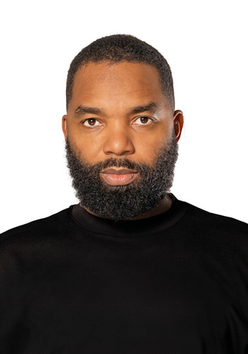 A man wearing a black turtleneck stares at the camera, with a serious expression against a white background