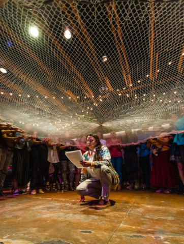 Carolina Caycedo crouches on the ground surrounded by a large group of people holding a net over her head