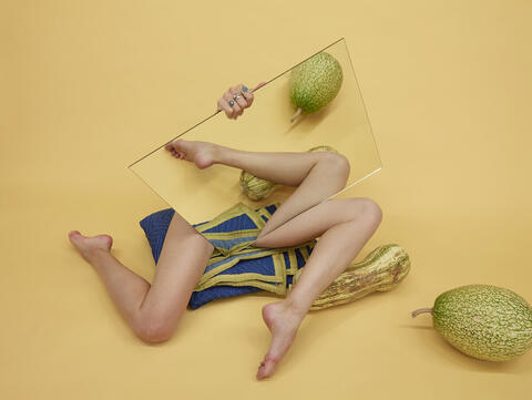 Two disembodied legs hidden by a mirror, surrounded by melons, on a yellow background