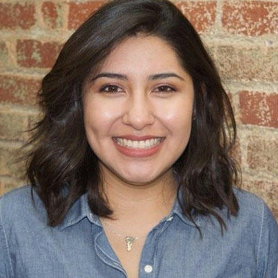 Portrait of Marisela Ramirez. Marisela is smiling and standing in front of a brick wall.