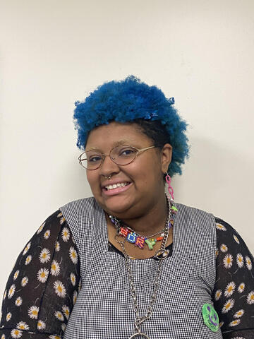 Portrait of Rory Hayes. They have blue hair and are smiling, looking into the camera, standing in front of a white background.