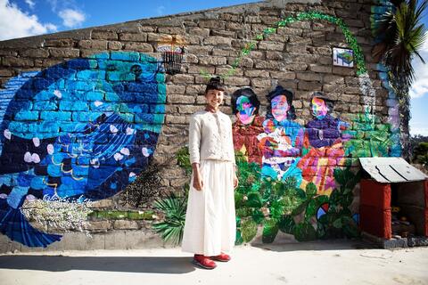 Artist iris yirei hu standing in front of a brick wall with several murals. 