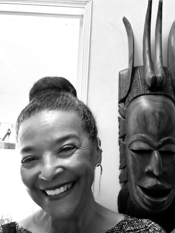 Black and white portrait of Sequoia standing in front of of a wall with a mask hanging on it.