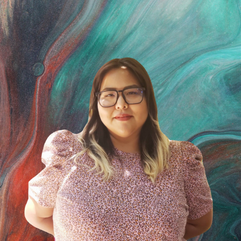 Portrait of Dria Yellowhair. Wearing a pink shirt and standing in front of a colorful background. 