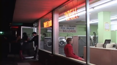 An exterior shot of a laundromat, with a woman wearing a red shirt visible through the window
