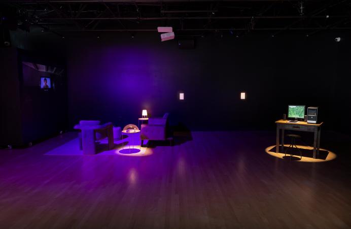 A living room setting bathed in purple light and a desktop computer beneath a spotlight, as part of the Encoding Futures exhibition