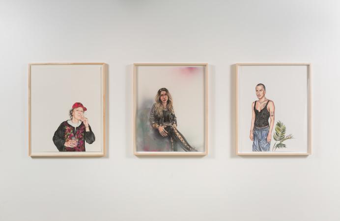 Three hand-drawn portraits, each on white and framed in light brown wood, side by side on a stark white wall