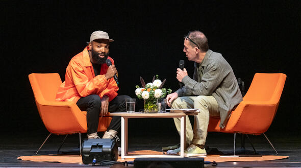 Stage view of Tavares Strachan and Paul Holdengräber sitting in orange chairs at angles across each other.