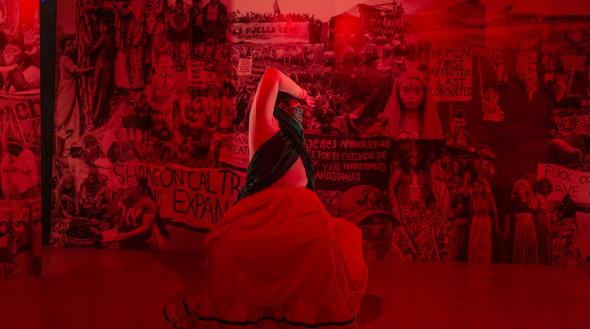 A woman lit in red bends backwards toward a wall covered in photos of people protesting